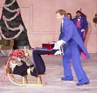 Drosselmeyer is pushing a sled with a doll on her back on the sled.  The doll's arms and legs are up in the air in a frozen position