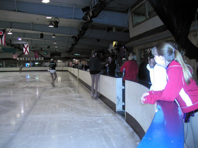 Skaters on the ice waiting to go on with spectaors watching from the chaperone area